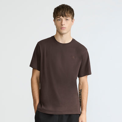A young man with short brown hair wears a Champion Classic T-Shirt featuring the C Logo and black pants. He stands against a light background, looking directly at the camera with a neutral expression.