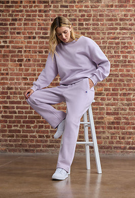 Woman in a light purple outfit sitting on a stool against a brick wall.